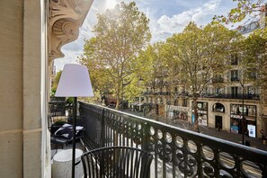 A balcony with a white table and chairs, overlooking a street with trees and buildings. - Superb New 1 Bedroom Flat Notre Dame de Paris (Paris)