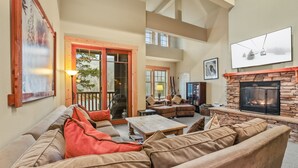 A spacious living room with a stone fireplace, comfortable seating, and a large window overlooking a deck. - Grandeur at Elevation 3 Bedroom Loft 3 Condo (Kirkwood)