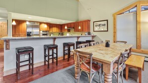 A modern kitchen with wooden cabinets, a central island with bar stools, and a dining table with chairs. - Grandeur at Elevation 3 Bedroom Loft 3 Condo (Kirkwood)