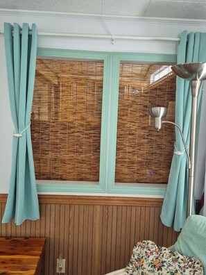 Une pièce avec une fenêtre recouverte de stores en bambou, un lampadaire et un coussin à motifs floraux posé sur un banc en bois.