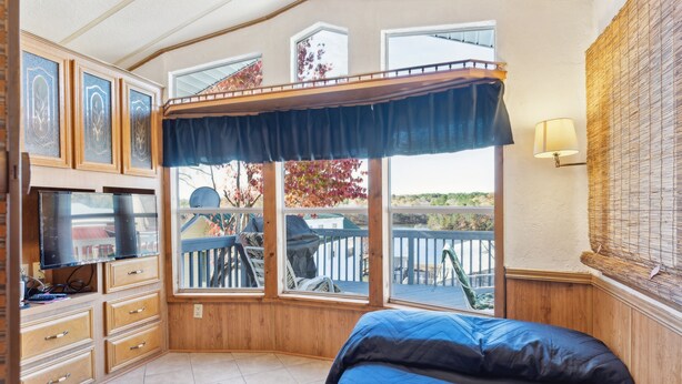 A bedroom with a bed, a TV, a desk with drawers, a window with a view of the outside, and a lamp.