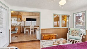 A cozy room with a wooden floor, a white bed, a wicker chair, and a small table.