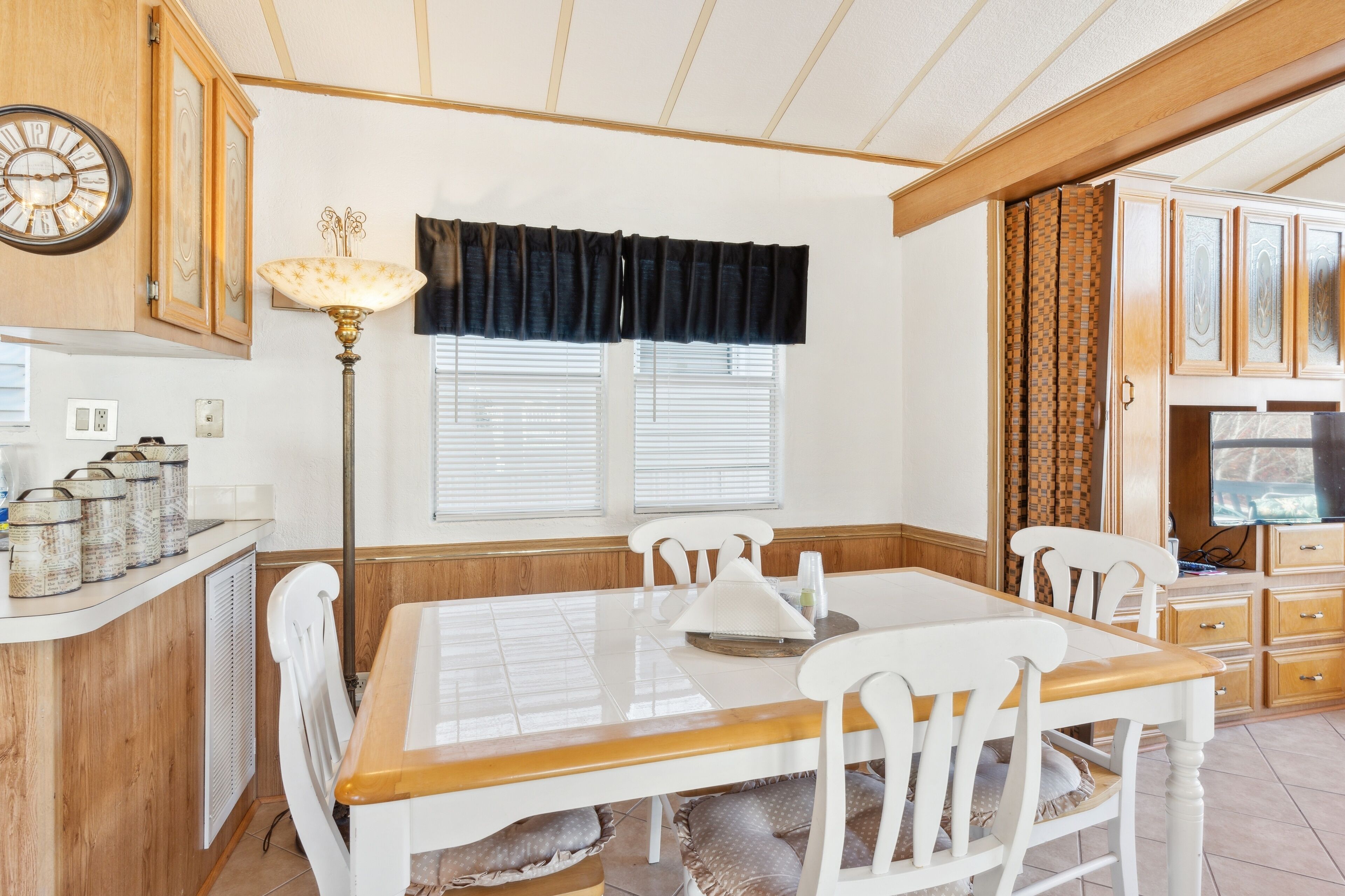 A dining area with a wooden table, white chairs, a clock, and a window with blinds.