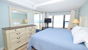Ein Schlafzimmer mit einem großen Bett, einem Schminktisch, einem Fernseher und einem Fenster mit Vorhängen.