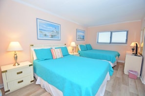 A hotel room with two beds, each with a teal bedspread and white bedding, a white nightstand with a lamp, a dresser with a mirror, and two framed pictures on the wall.