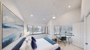 Une chambre moderne avec un grand lit, un bureau et une chaise. De grandes fenêtres offrent une vue sur les arbres et un immeuble.