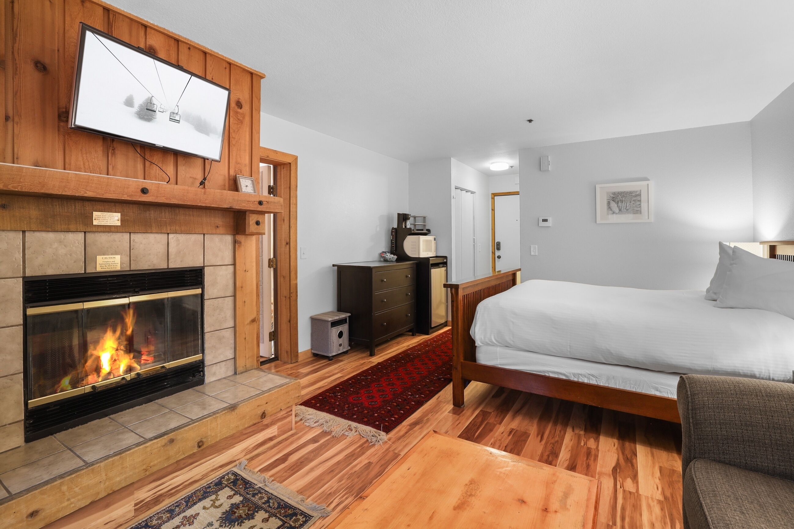 A bedroom with a fireplace, a bed, a dresser, and a television mounted on the wall.