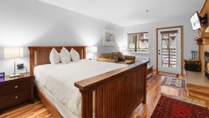 A bedroom with a large bed, a wooden headboard, a nightstand with a lamp, a sofa, a fireplace, and a view of the outside through a door. - Kirkwood Retreat - 2 Bedroom Condo 2 Condo (Kirkwood)