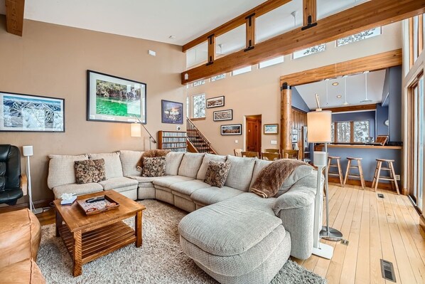 A spacious living room with a large sectional sofa, a wooden coffee table, and a fireplace. There are multiple framed pictures on the walls, a staircase leading to an upper level, and a kitchen area visible through an open doorway. - 2997 Basingdale 3 Bedroom Home (Vail)
