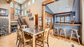 Un'ampia zona giorno con soffitto alto, travi in legno, un tavolo da pranzo con sedie, una cucina con bancone e una scala che conduce a un piano superiore.
