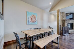 A dining area with a wooden table and chairs, a painting of pancakes on the wall, and a stone fireplace in the background. - Luxury Home - Sleeps 10 - Fireplace & Mountain View 3 Bedroom Condo (Keystone)