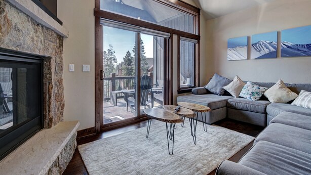 A modern living room with a stone fireplace, large sliding glass doors to a deck, a gray sectional sofa, and three wall-mounted paintings. - Luxury Home - Sleeps 10 - Fireplace & Mountain View 3 Bedroom Condo (Keystone)