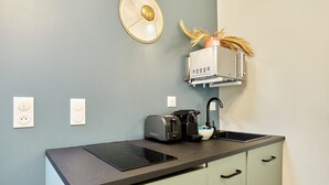 A compact kitchenette with a black induction cooktop, a toaster, and a coffee maker. There are two wall-mounted outlets and a small shelf with a decorative item.