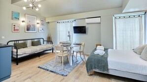 A compact, well-lit room with a bed, a sofa, a dining table with chairs, and a TV mounted on the wall.