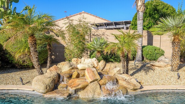 Piscina com cachoeira de pedra, cercada por palmeiras e um edifício com telhado inclinado.