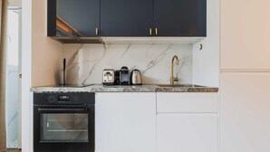 Una cocina compacta con armarios oscuros, horno integrado, un lavamanos con grifo dorado y encimera de mármol.