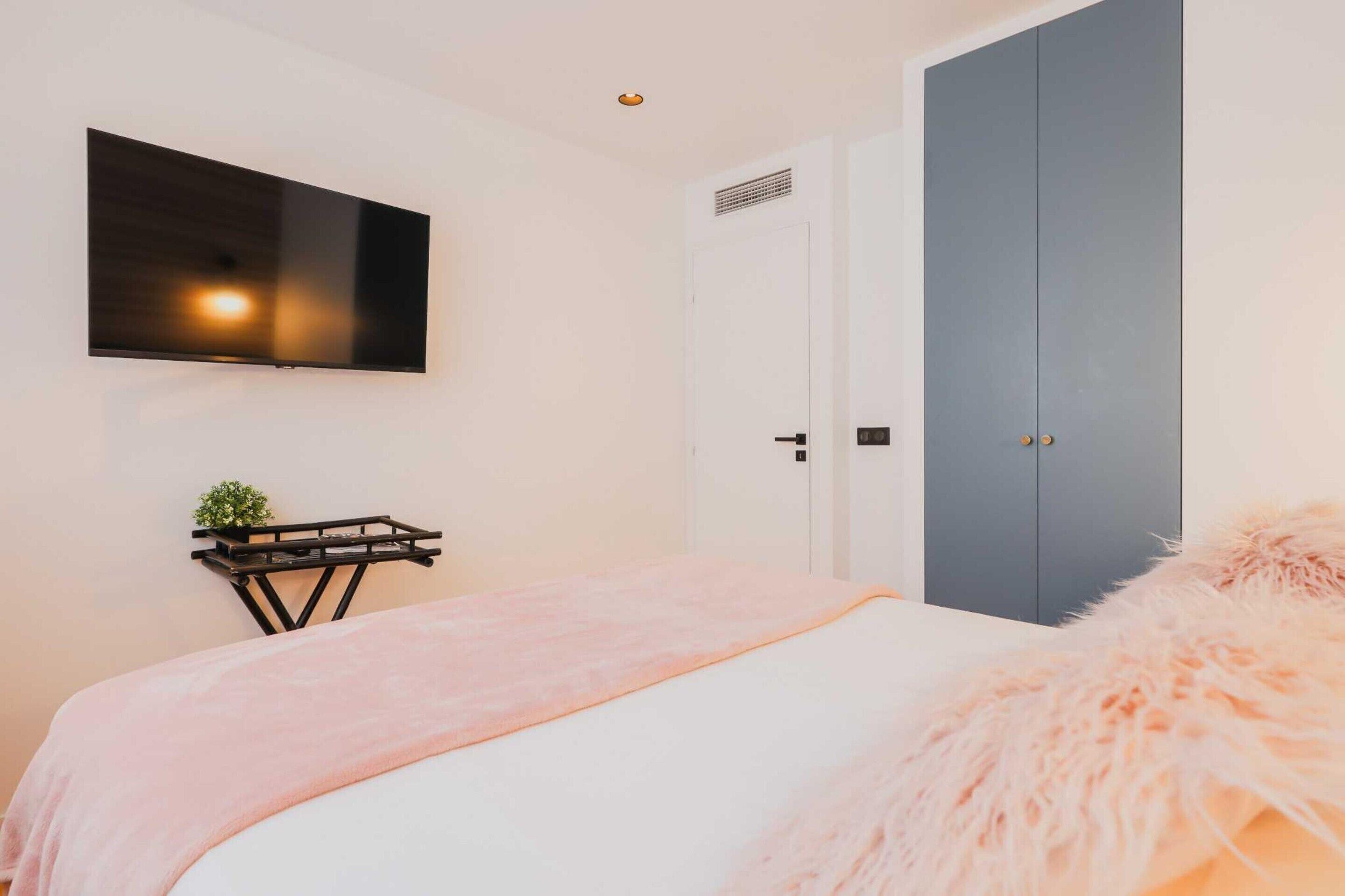 Une chambre à coucher avec un lit, un téléviseur fixé au mur, une armoire et une petite table sur laquelle est posée une plante.