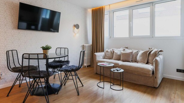 Una sala de estar moderna con un televisor de pantalla plana montado en una pared texturizada, un sofá beige, una mesa de centro redonda y un comedor con una mesa y sillas de estructura metálica negra.