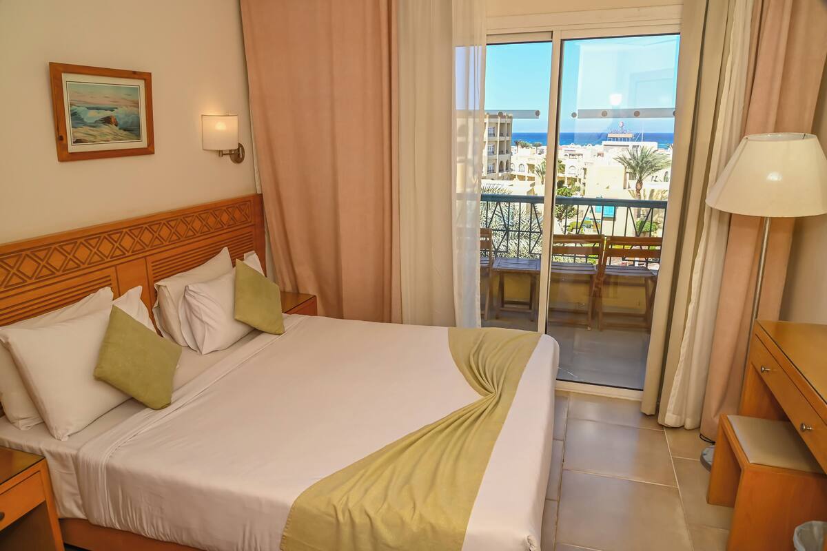 A hotel room with a bed, a nightstand, a lamp, a painting, and a balcony with a view of buildings and the sea.