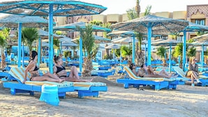 A beachside resort with multiple sun loungers under blue umbrellas, palm trees, and a sandy beach. - Eagles Downtown Zahabia Aqua Park (Hurghada)