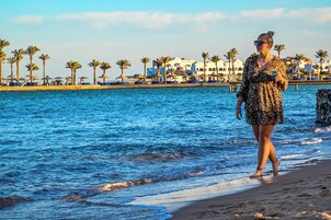 A beach with a woman walking along the shore, palm trees, and a building with a white facade in the background.