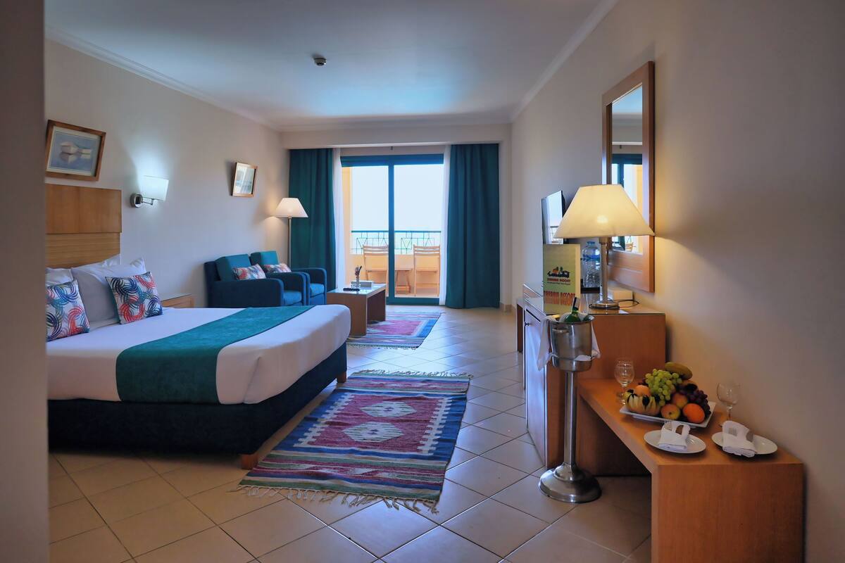 A hotel room with a bed, a desk with a lamp, a chair, a small table, and a balcony view.