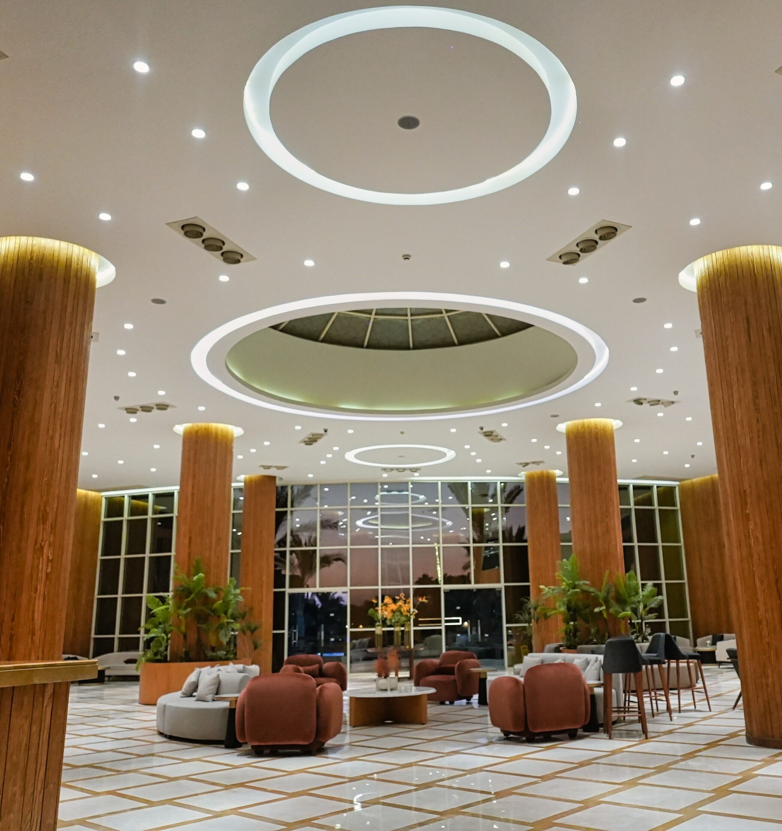 A spacious lobby with a modern design, featuring a central seating area with sofas and chairs, large wooden columns, and a patterned floor.