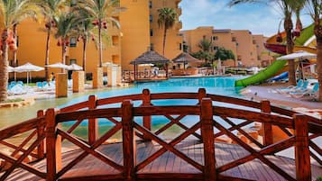 A resort pool area with a wooden bridge, palm trees, and a water slide.