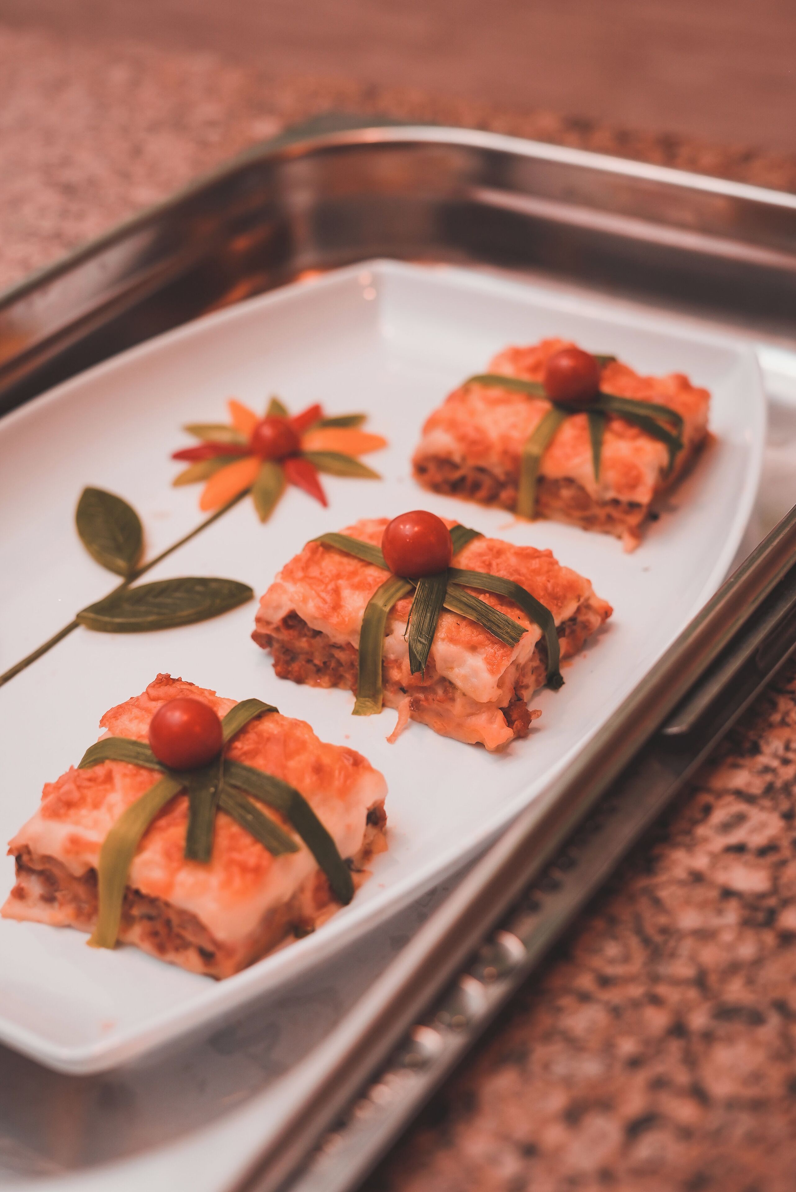 Une assiette rectangulaire blanche contenant trois portions de nourriture, décorées de feuilles vertes et de baies rouges, est posée sur un comptoir en granit.