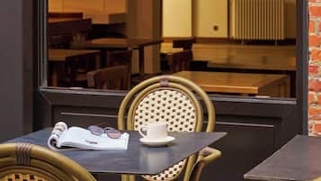 A table with a book and glasses, a wicker chair, and a window with a view of indoor seating.