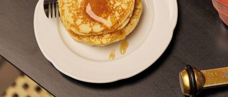 A breakfast setting with pancakes on a plate, a cup of coffee, and a bowl of cereal with fruits.