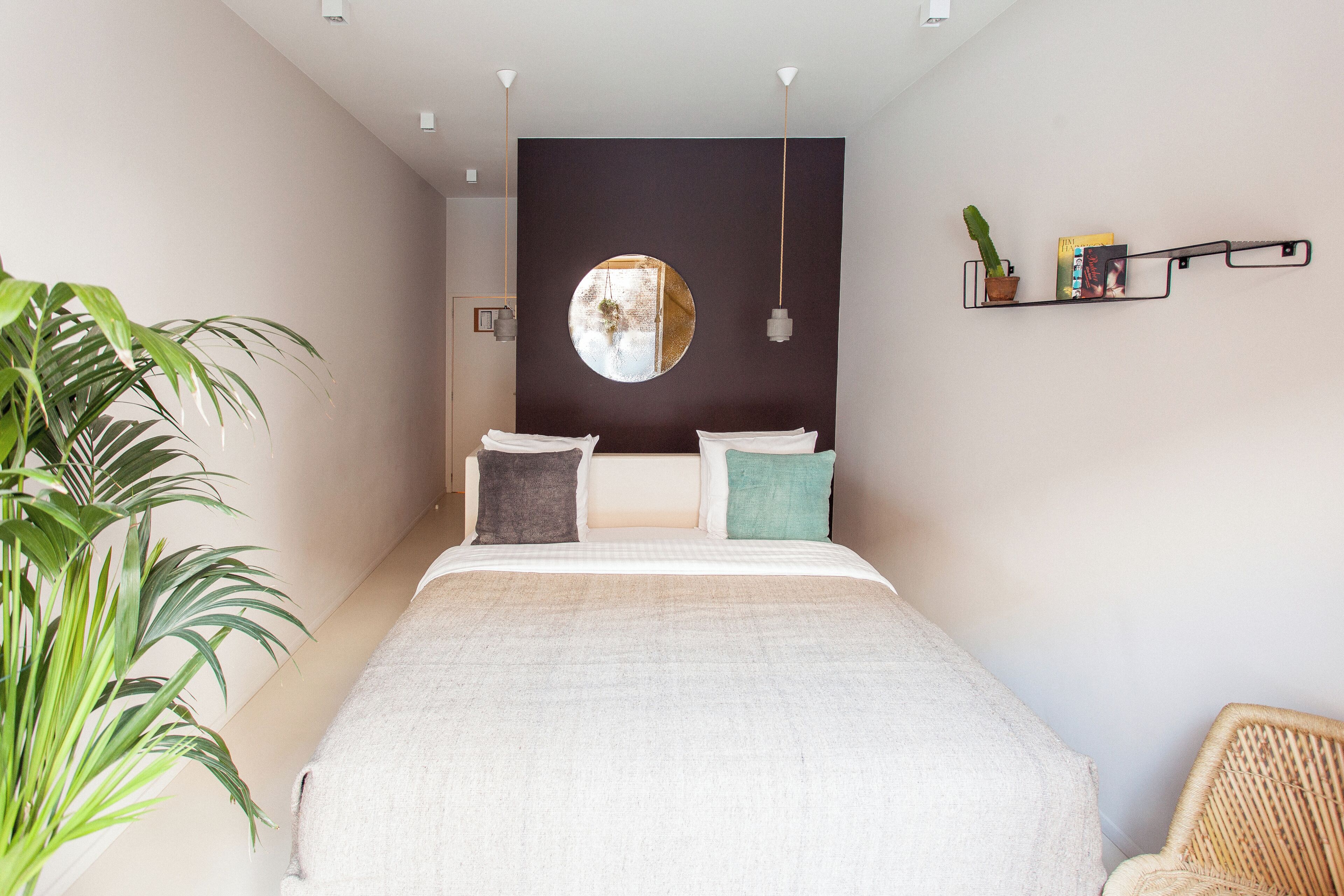 Un dormitorio con cama, almohadas, un espejo redondo, una lámpara colgante, una planta en maceta y una estantería con libros y un cactus.