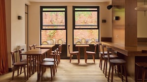 Comedor con mesas y sillas de madera, ventanas amplias con vistas a una pared de ladrillo y una lámpara moderna.