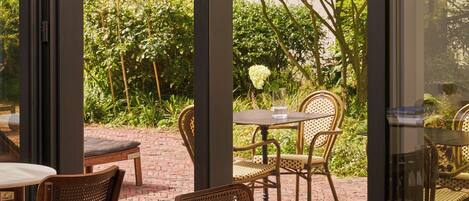 An outdoor seating area with tables and chairs, a brick pathway, and a view of a garden through large windows.