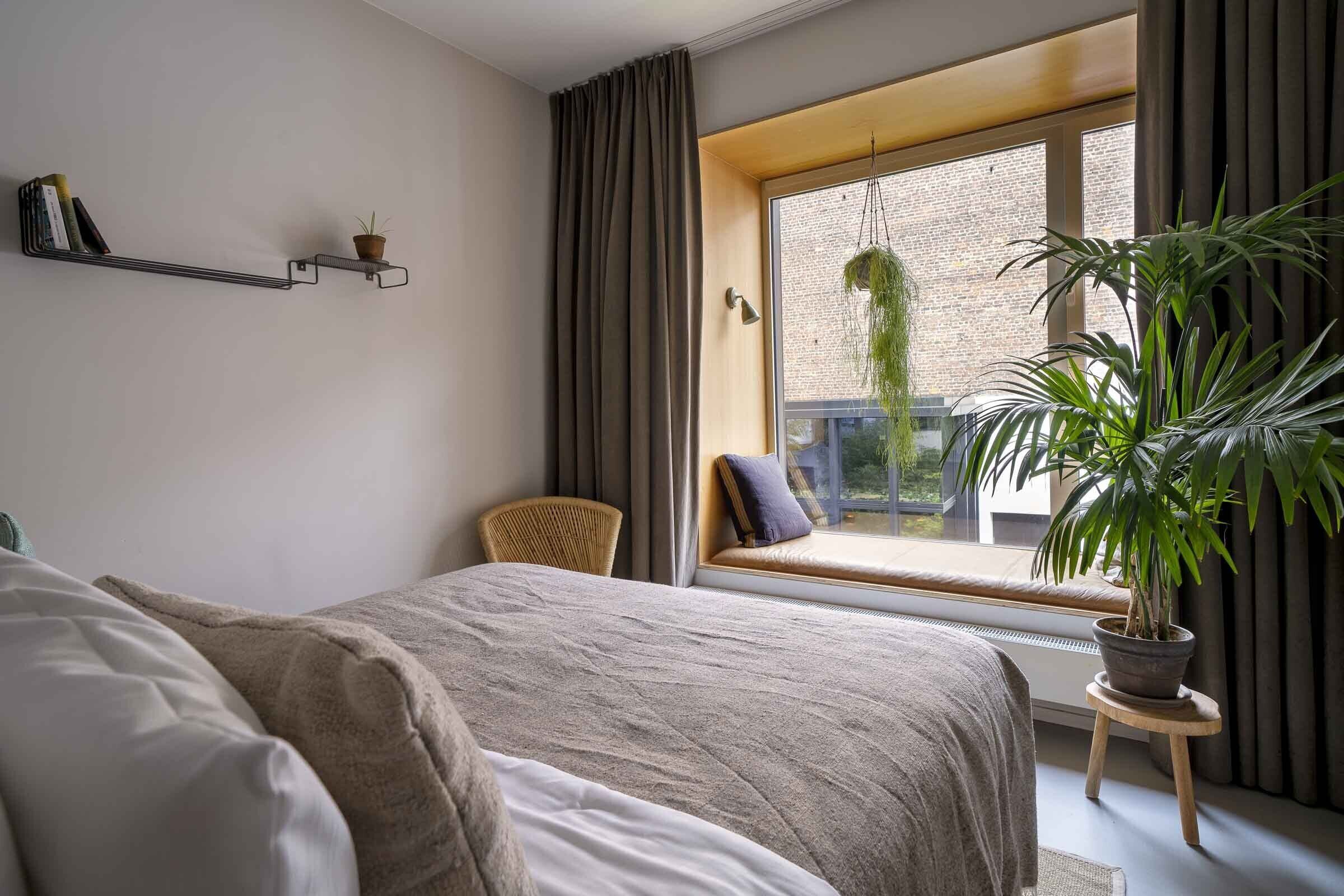 Un dormitorio con cama, un asiento junto a la ventana, una planta en maceta y una estantería con libros y una planta.