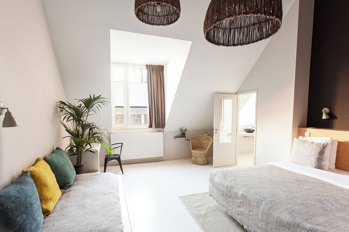 A modern bedroom with a large bed, a sofa, a potted plant, and a wicker chair.