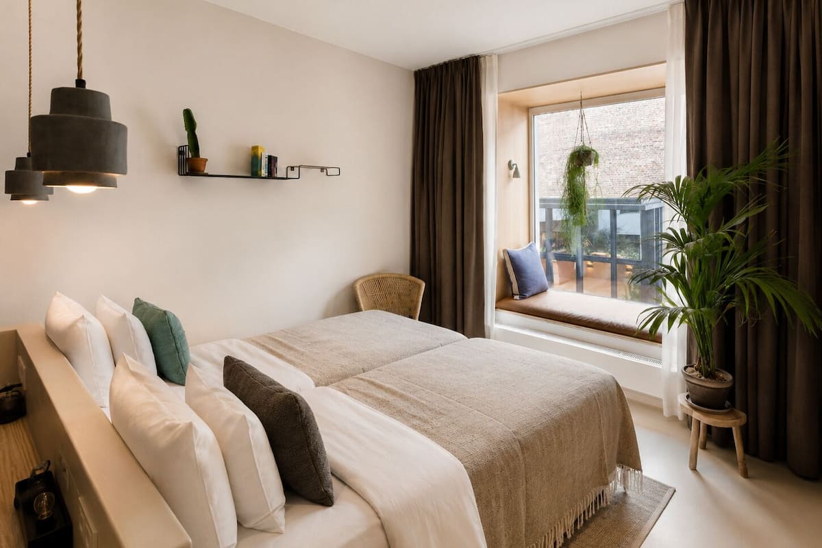 A modern bedroom with a large bed, a hanging light fixture, a window seat with a view, and a potted plant.