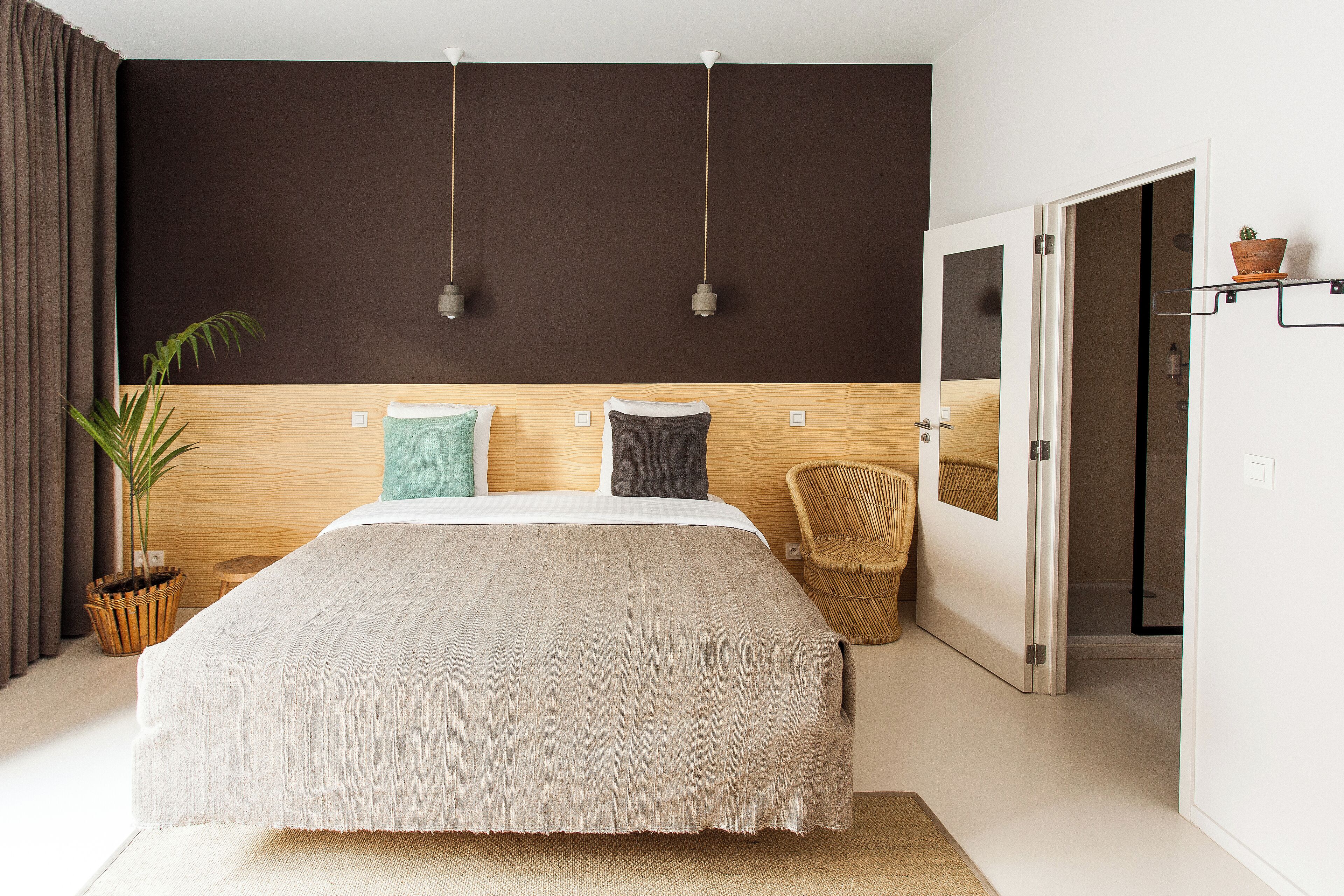 Un dormitorio con una cama grande, una planta en maceta, un sillón de mimbre y una puerta que da a un baño.