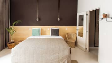 Un dormitorio con una cama grande, una planta en maceta, un sillón de mimbre y una puerta que da a un baño.