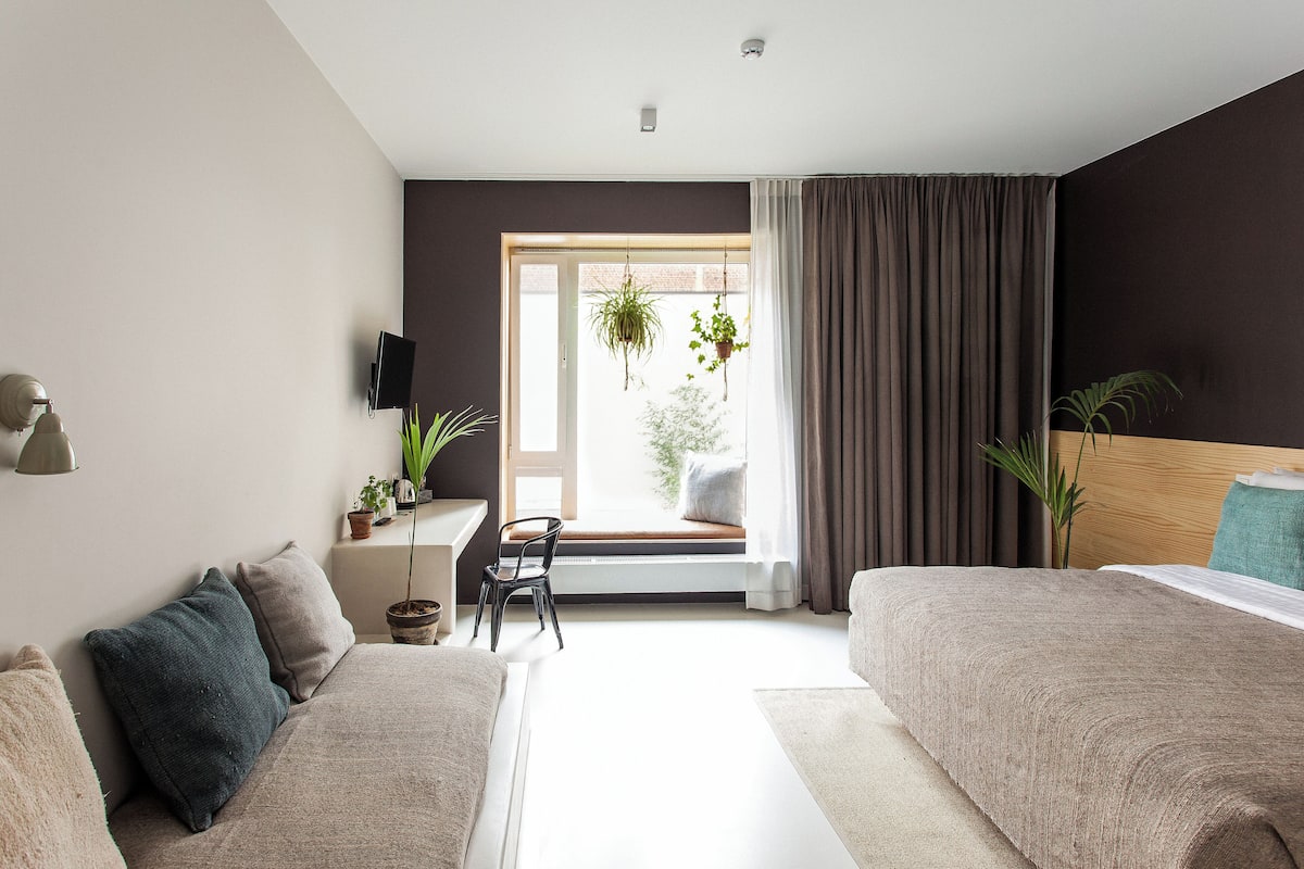 A modern bedroom with two beds, a desk with a chair, and a large window with curtains.