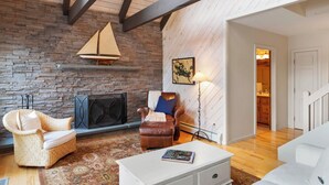 Una sala de estar acogedora con una chimenea de piedra, un sillón de cuero marrón, un sillón de mimbre y un sofá blanco con una mesa de centro blanca.