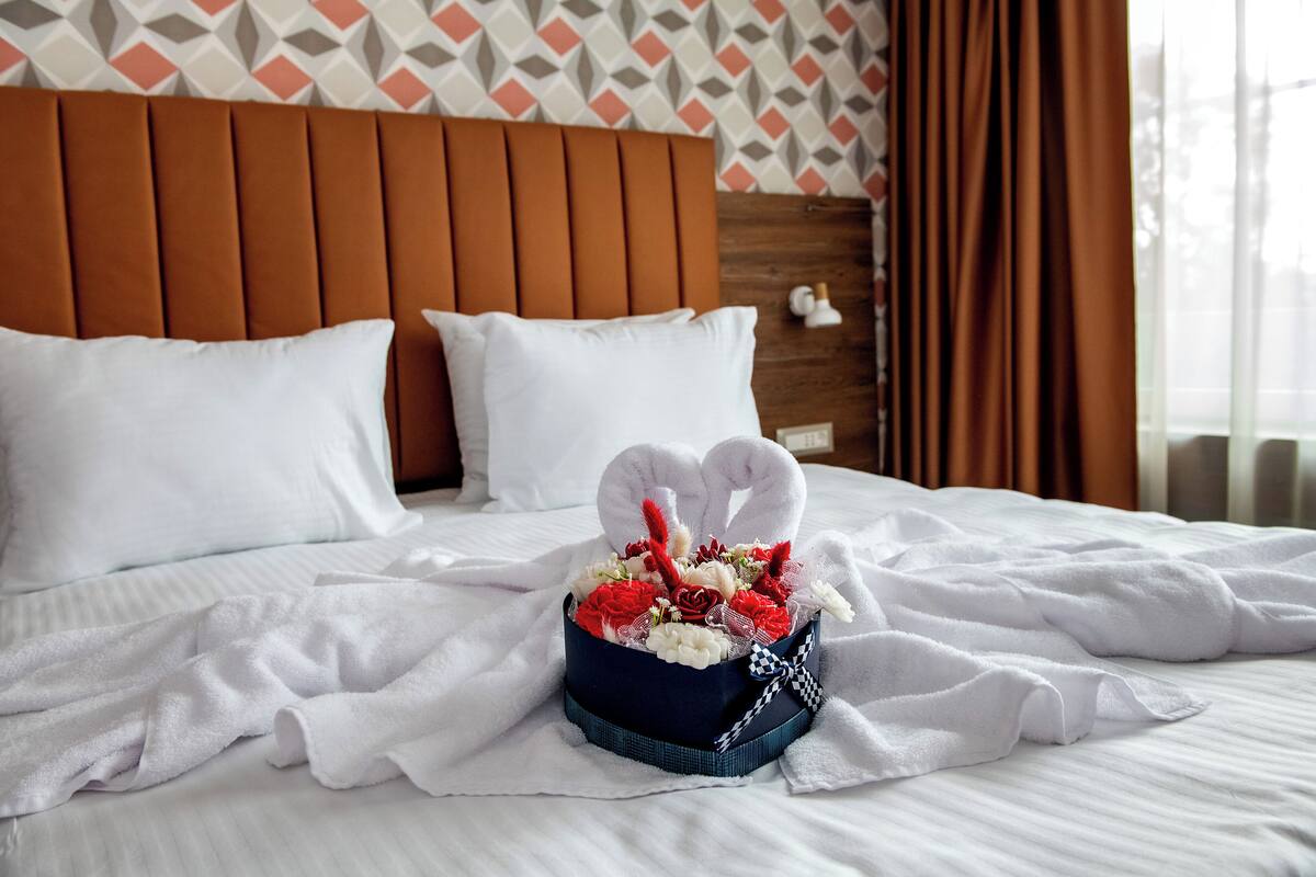 A neatly made bed with white linens, a heart-shaped floral arrangement in a blue box, and a wooden headboard.