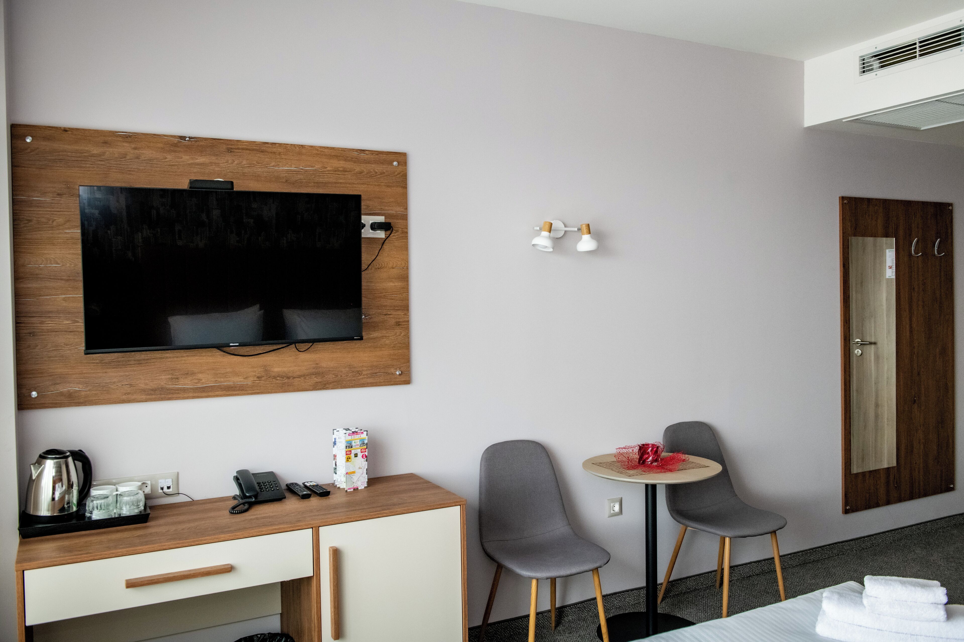 Habitación de hotel con un escritorio de madera, un televisor montado en la pared, una mesita redonda con dos sillas, un minibar y una puerta con espejo.