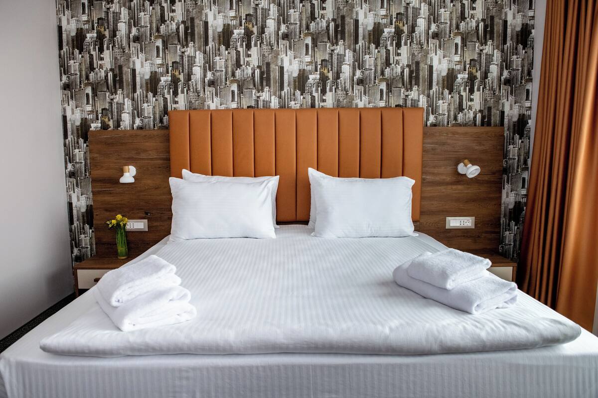A neatly made bed with white linens and pillows, a wooden headboard, and a cityscape wallpaper.