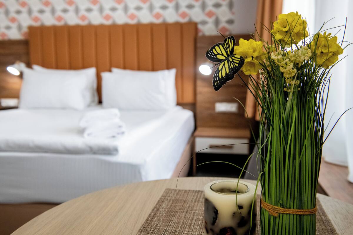 A hotel room with a neatly made bed, a bedside table with a candle, and a vase with yellow flowers and a butterfly.