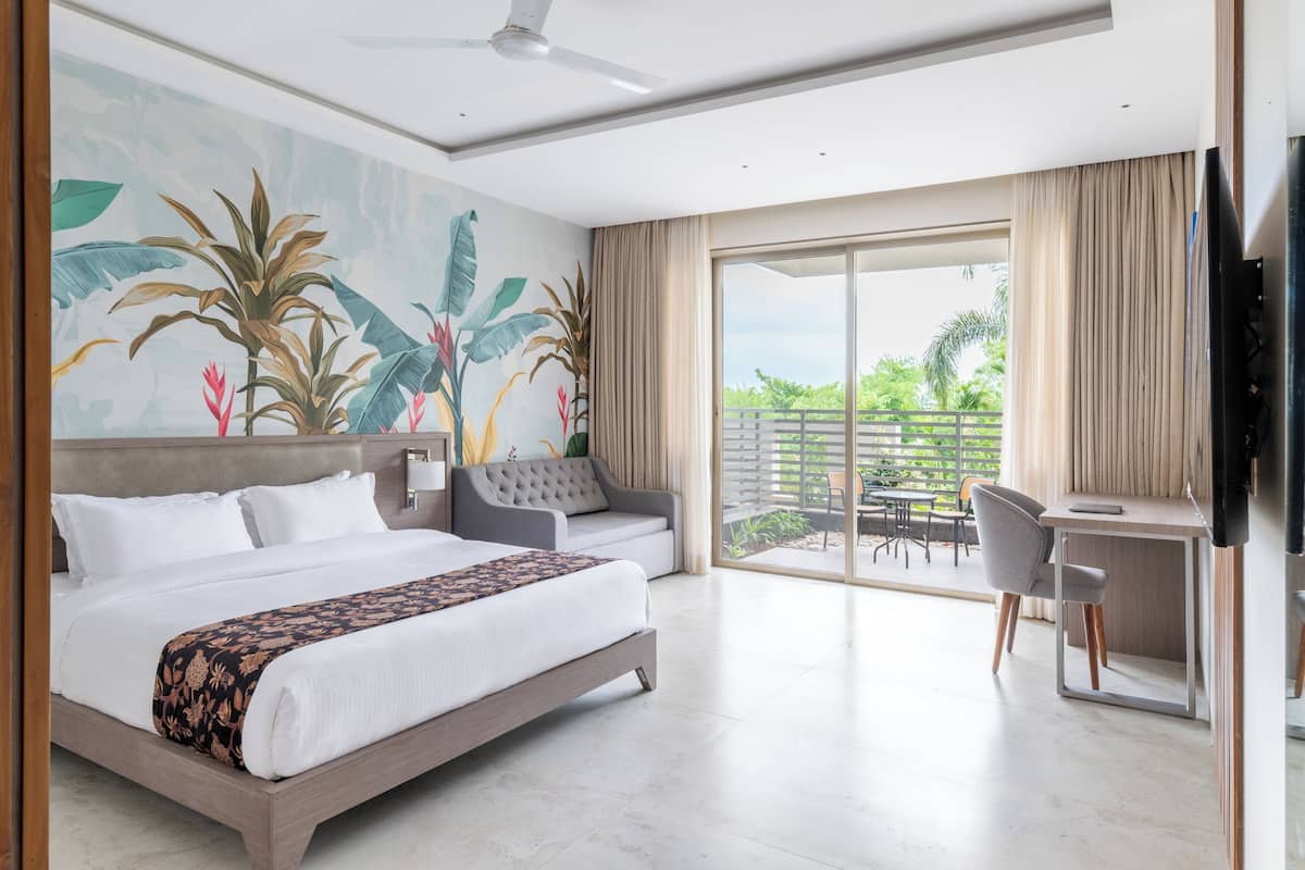 A hotel room with a large bed, a desk, and a balcony with a view of greenery.