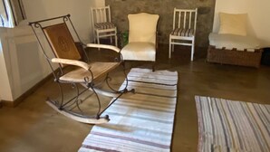 Ein gemütliches Wohnzimmer mit einer Steinwand, einer Holzbalkendecke und einem Steinkamin. Es gibt zwei weiße Sessel, einen Schaukelstuhl und ein kleines Bett mit Kissen.
