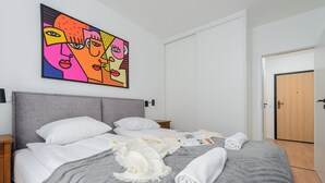 A neatly made bed with white linens and a gray blanket, a wooden nightstand, a wall-mounted painting, and a wooden door.