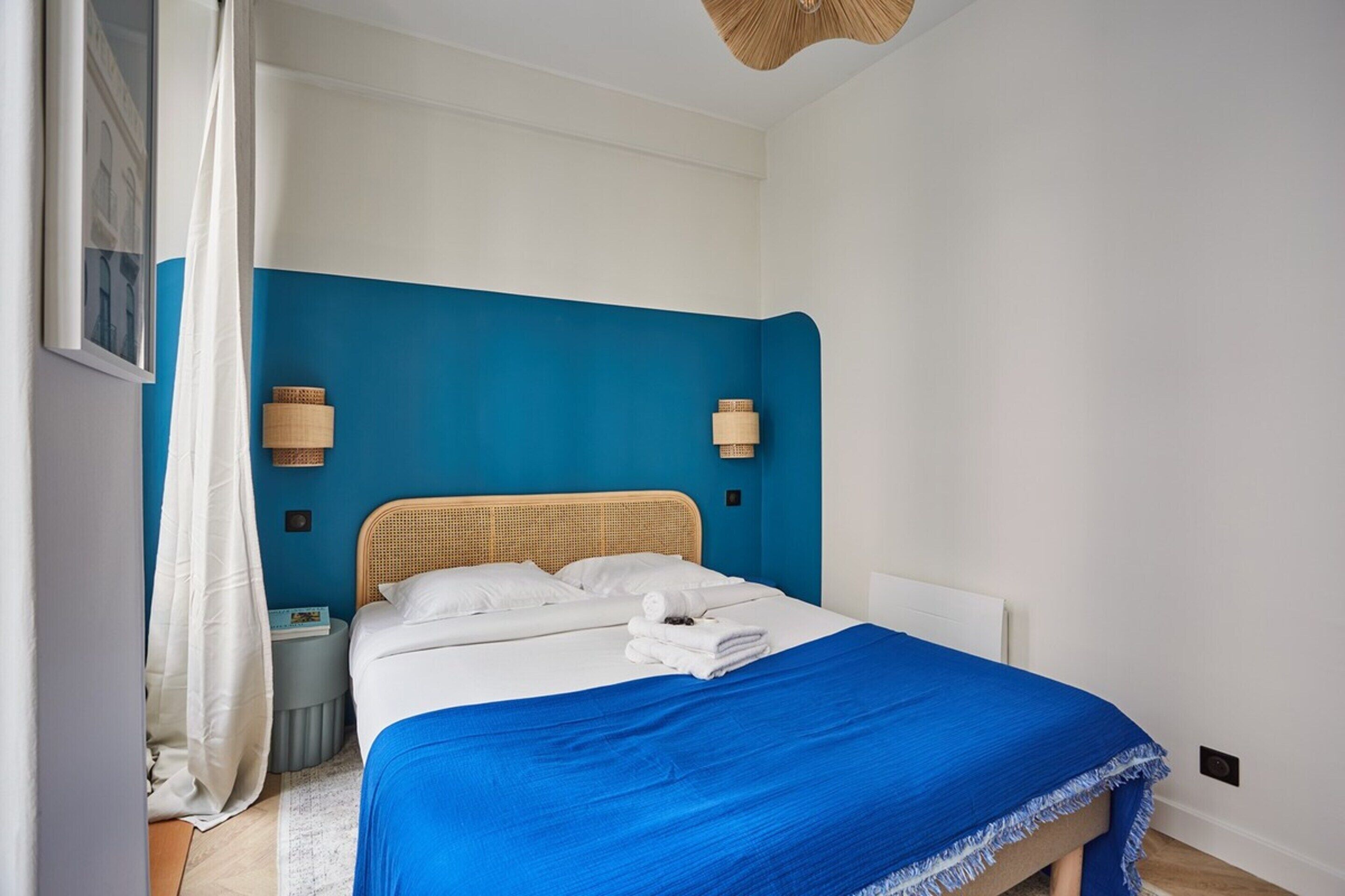 A neatly made bed with a blue bedspread, white pillows, and a white headboard. There are two wall-mounted lamps on either side of the bed.