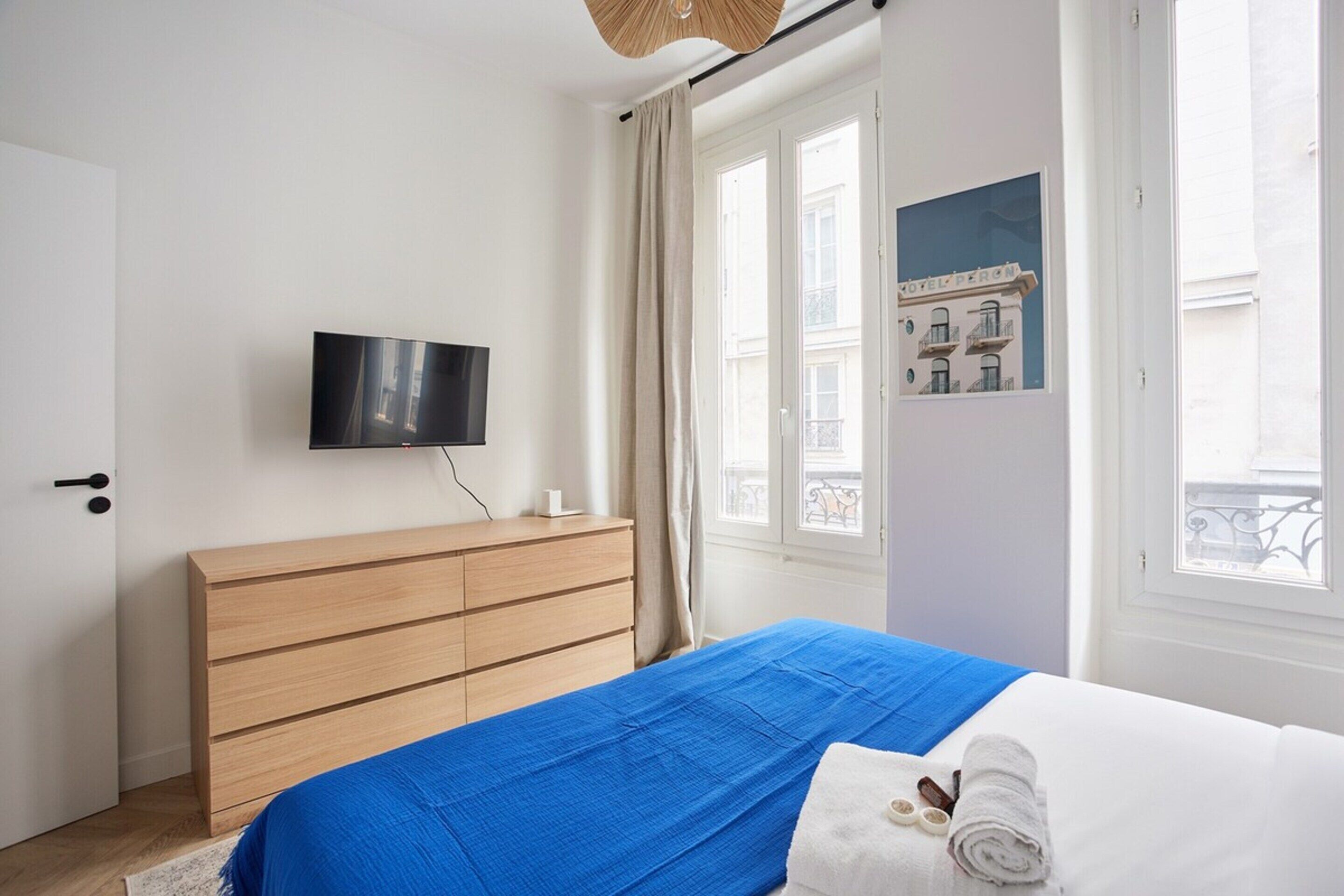 A bedroom with a bed, a wooden dresser, a flat-screen TV, and a window with a view of buildings.